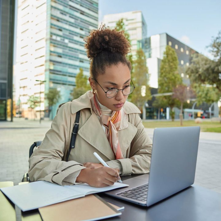 Busy student prepares for university admission watches training webinar concentrated in laptop screen wears transparent glasses and coat poses at desk outdoors. Businesswoman works online at city