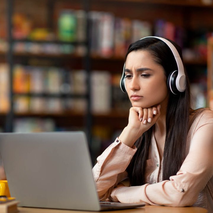 Online education concept. Concentrated latin woman studying online at cafe, using laptop and headset, copy space. Arab female student having online lesson, looking at laptop screen