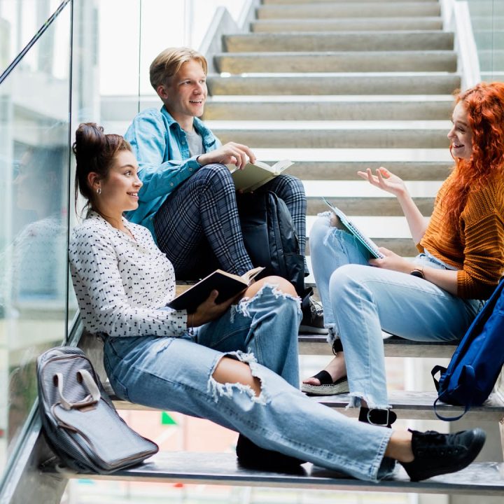 Cheerful university students sitting on stairs and talking indoors, back to school concept.